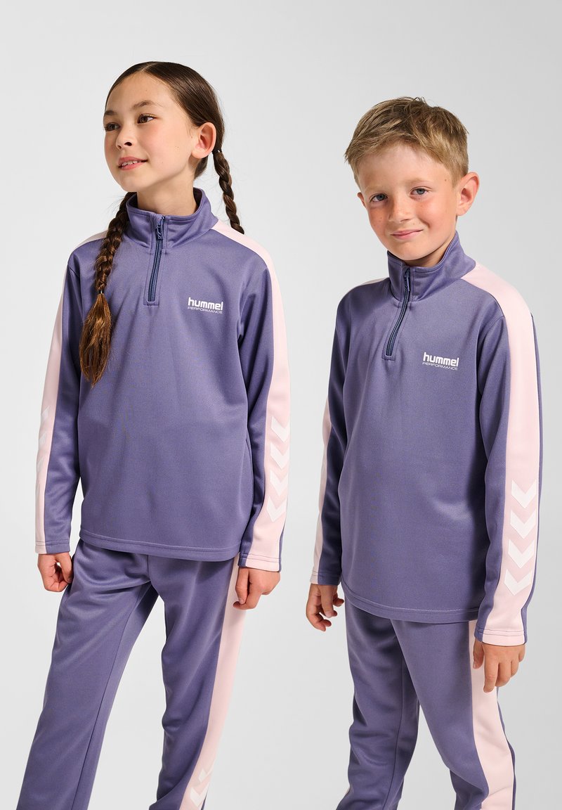 Lilla træningssæt med pink detaljer, med lynlås ved kraven og chevronmønster på ærmerne. Fremstillet af glat, elastisk materiale.