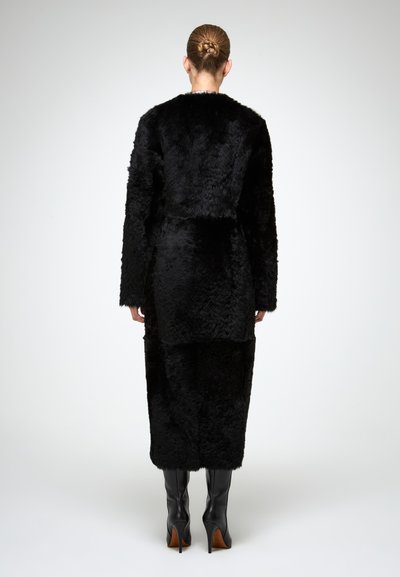 Cappotto di pelliccia finta nera con silhouette allungata, maniche lunghe e superficie texture. Indossato con stivali alti fino al ginocchio neri con tacco a stiletto.