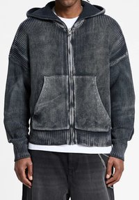 Felpa grigia con cappuccio, zip e texture a costine, tasca frontale e un leggero sbiadimento. Presenta una cerniera a contrasto lungo il centro.