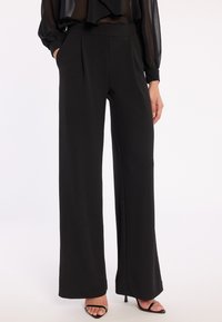 Zwarte wijde broek van soepel stof, met plooien op de taille en zijzakken, gecombineerd met een doorschijnende zwarte blouse en hoge hakken.