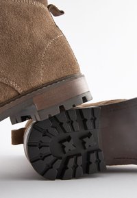 Hnedé semišové členkové topánky s textúrovanou čiernou gumovou podrážkou a robustným vzorom trakcie; majú bočný slučku pre jednoduché obúvanie.