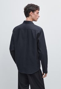 Schwarzes Langarmhemd aus glattem Stoff mit klassischem Kragen, geknöpften Manschetten und abgerundetem Saum, von hinten gezeigt.