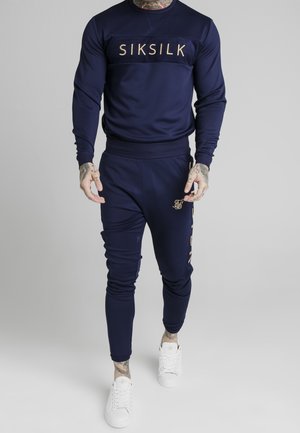 Mężczyzna w granatowej bluzie SikSilk i dopasowanych spodniach dresowych, połączonych z białymi sneakersami, stojący na jednolitym szarym tle.