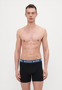 Schwarze Boxershorts mit einem blauen Bund, auf dem "BJÖRN BORG" steht. Der Stoff wirkt glatt und hat eine enganliegende Passform.