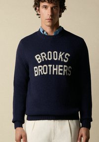 Maglione a maglia blu navy con la scritta "BROOKS BROTHERS" in lettere bianche. Polsini e orlo a coste, indossato sopra una camicia celeste con colletto.