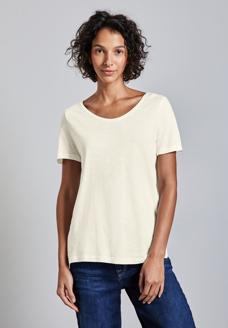 Lys beige kortærmet T-shirt med rund halsudskæring og afslappet pasform, parret med mørkeblå jeans. Blødt, let tekstureret stof.