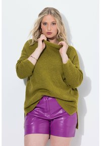 Maglione oversized in verde oliva con collo alto e spalle scese, abbinato a pantaloni corti attillati e lucidi di colore viola. Tessuto texturizzato, vestibilità rilassata.