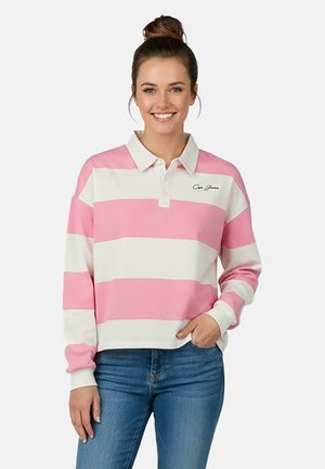 Vrouw met haar in een knot, gekleed in een roze-wit gestreept poloshirt met lange mouwen en een blauwe spijkerbroek, glimlachend met één hand in haar zak.