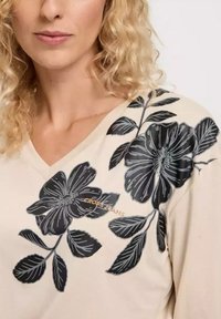 Beżowa bluzka z dekoltem w serek z czarnym kwiatowym wzorem na jednym ramieniu. Charakteryzuje się miękką teksturą i subtelnym logo marki na klatce piersiowej.