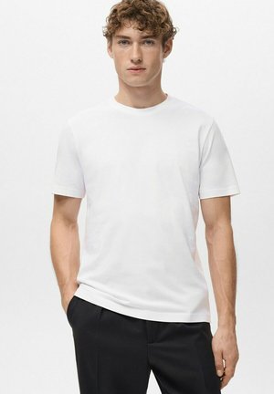 Junger Mann mit lockigem Haar, der ein schlichtes weißes kurzärmliges T-Shirt und schwarze Hosen trägt, steht mit einer Hand in der Tasche.