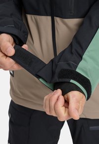 Technical winter jacket featuring a three-color design: black, beige, and green. Includes Velcro cuffs, textured fabric, and zippered pockets.
