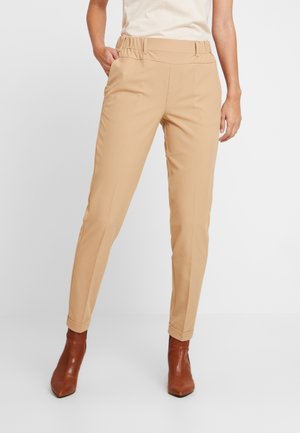 Pantalons beige ajustés avec une large ceinture et des plis, dotés d'un tissu lisse et d'une jambe effilée, associés à des bottines à talons bruns.