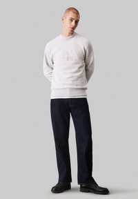 Suéter gris claro con logo en relieve, cuello redondo y puños acanalados, combinado con jeans rectos de color azul oscuro y zapatos negros.