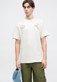 Ανοιχτό μπεζ βαμβακερό μπλουζάκι με κοντά μανίκια, που διαθέτει ροζ γραφικά και κείμενο. Συνδυασμένο με ελαιόχρωμα παντελόνια και κρατώντας ένα μπλε καπέλο.