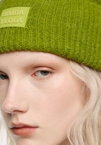 Gorro de punto verde con textura acanalada y una etiqueta rectangular con el logo "BIMBA Y LOLA" en verde claro. Primer plano del rostro del modelo.