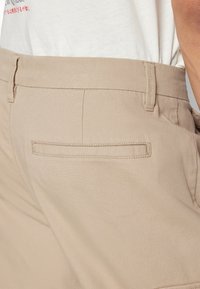 Beige broeken met een gladde textuur, een platte achterzak en een strakke tailleband. De stof lijkt kreukbestendig en duurzaam te zijn.