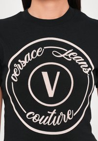 Juodas marškinėliai su dideliu šviesiai pilku logotipu, turinčiu apvalios formos dizainą. Ryškiai matomas tekstas "Versace Jeans Couture" ir "V" emblema.