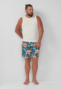 Witte mouwloze shirt en blauwe zwemshorts met bloemen in levendige oranje en roze kleuren tegen groene bladeren; blote voeten.