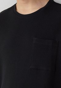 Czarny t-shirt z dzianiny waffle z okrągłym dekoltem i małą kwadratową kieszonką na piersi. Tekstura jest ryflowana, a krój dopasowany.