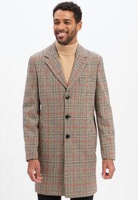 Lächelnder Mann in einem beigen Rollkragenpullover und einem karierter beigen, schwarzen und roten langen Mantel mit drei schwarzen Knöpfen, der vor einem weißen Hintergrund steht.