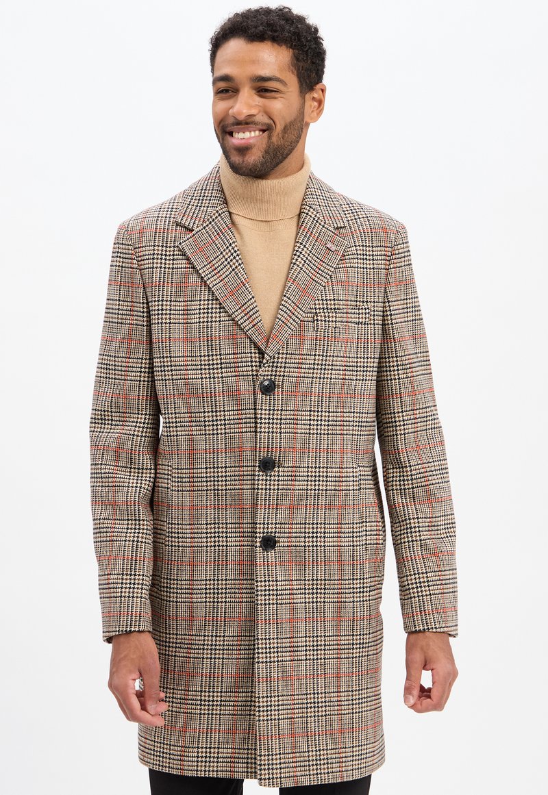 Lächelnder Mann in einem beigen Rollkragenpullover und einem karierter beigen, schwarzen und roten langen Mantel mit drei schwarzen Knöpfen, der vor einem weißen Hintergrund steht.