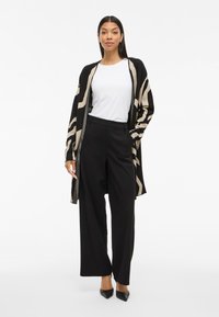 Γυναίκα που φοράει ένα μαύρο και μπεζ καρό κασκόλ πάνω από ένα λευκό πουκάμισο, μαύρες παντελόνες φαρδιές και μαύρα παπούτσια με μυτερή μύτη, στέκεται με αυτοπεποίθηση.