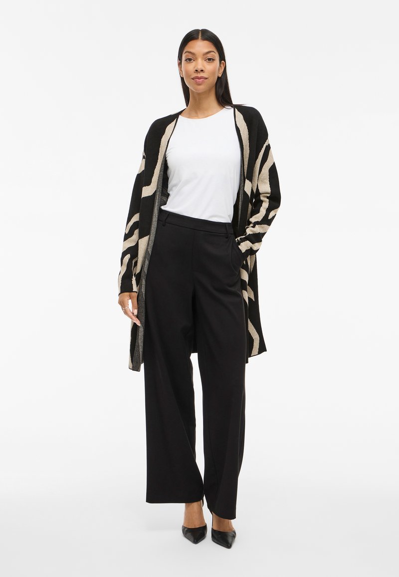 Γυναίκα που φοράει ένα μαύρο και μπεζ καρό κασκόλ πάνω από ένα λευκό πουκάμισο, μαύρες παντελόνες φαρδιές και μαύρα παπούτσια με μυτερή μύτη, στέκεται με αυτοπεποίθηση.