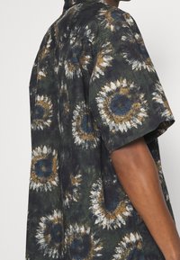 Camicia a maniche corte in tessuto scuro con motivo di girasoli in bianco, giallo e blu, dal taglio rilassato e dalla consistenza morbida.