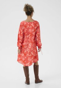 Blumenkleid in Koralle mit Puffärmeln, elastischen Bündchen und lockerer Passform, kombiniert mit braunen kniehohen Stiefeln.