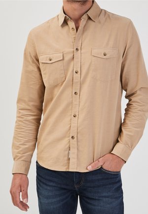 Mężczyzna w beżowej koszuli na guziki z dwoma kieszeniami na piersi i ciemnoniebieskich dżinsach, z jedną ręką w kieszeni, na jednolitym tle.