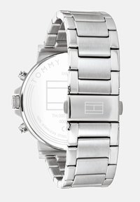 Montre en acier inoxydable argenté avec un bracelet maillé, boîtier rond et verre transparent mettant en valeur le logo de la marque et les détails au dos.