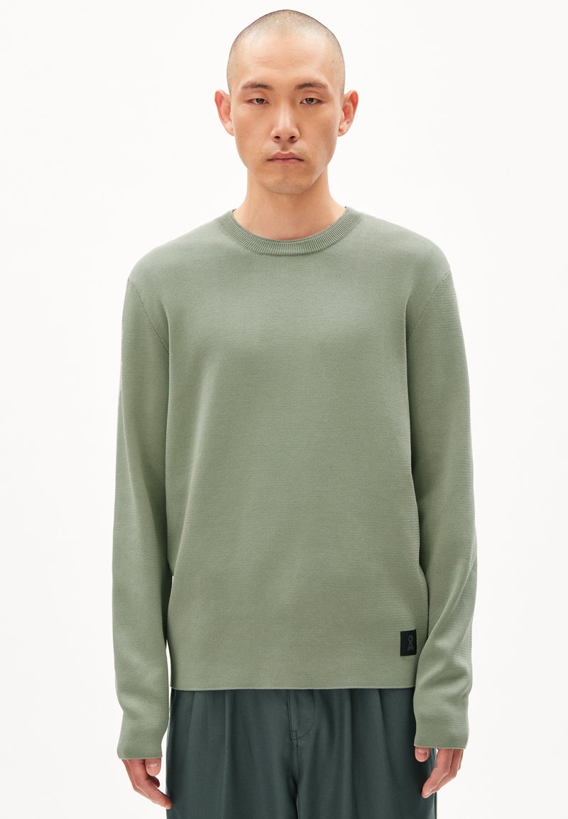 Suéter de manga larga verde claro en una textura de punto acanalado, con cuello redondo y un pequeño parche de logo negro en el dobladillo.