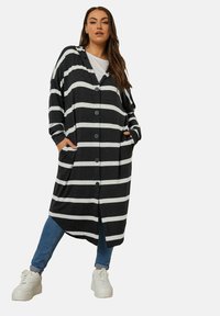 Mustavalkoinen raidallinen neuletakki, jossa on napillinen edusta, yliaistettava istuvuus, pitkät hihat ja sivutaskut. Päällä harmaan värinen T-paita.