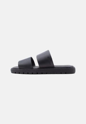 Crna ravna natikača s dva široka teksturirana remena i debelim potplatom s profilom, prikazana na jednobojnoj pozadini.