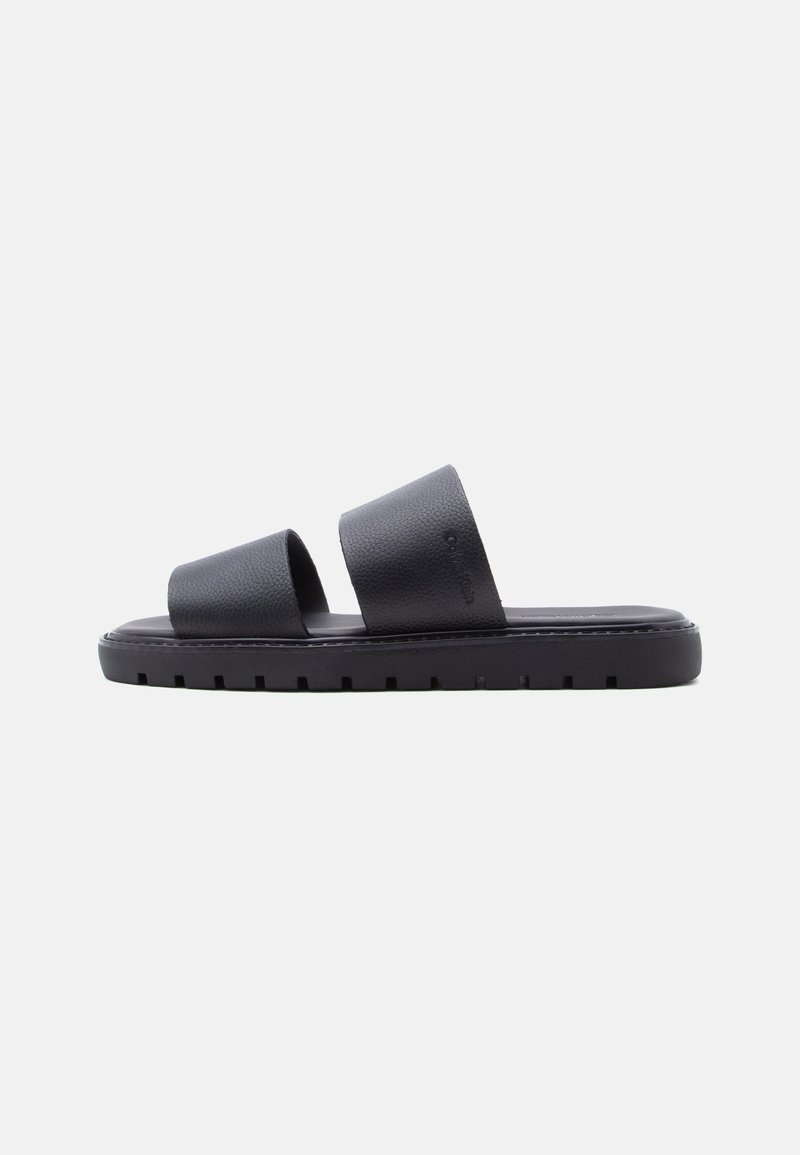 Sandale plate noire avec deux larges brides texturées et une semelle épaisse à crampons, présentée sur un fond uni.