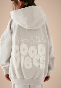 Ljusgrå överdimensionerad hoodie med en plyschliknande textur och en kenguruficka; vit "GOOD VIBES" grafik på baksidan och "Design Studio" text ovanför.