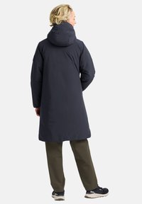 Lange marineblauwe pufferjas met een aansluitende capuchon, tekstuur oppervlak, losse pasvorm en zijzakken; gecombineerd met olijfgroene broek en sneakers.
