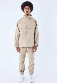Chaqueta impermeable beige con capucha y dos bolsillos frontales, combinada con pantalones cargo a juego y zapatillas blancas, con puños elásticos.