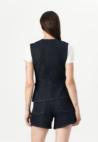 Donkere denimvest met een slim ontwerp en ronde halslijn. De achterkant heeft verticale naden. Gecombineerd met denimshorts, wat een casual stijl laat zien.