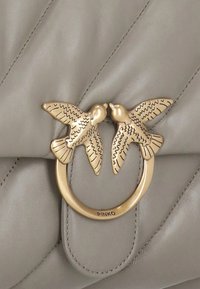 Chiusura in metallo dorato a forma di due uccelli che si affrontano su uno sfondo di pelle beige chiaro trapuntata, con "PINKO" inciso sull'anello.