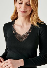 Top nero a maniche lunghe con scollatura a V profonda, decorato con dettagli in pizzo nero intricato, che mette in risalto motivi floreali e un materiale testurizzato.