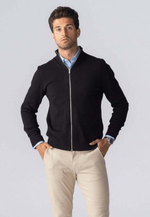 Sort zip-up sweater i blødt stof med ribbede manchetter og kant, parret med en lyseblå skjorte og beige chinos, vist på en ensfarvet baggrund.