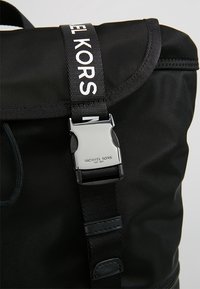 Sac Michael Kors noir avec boucle argentée et bandoulière noire imprimée de « MICHAEL KORS » en lettres blanches.