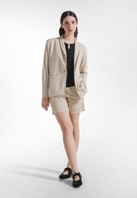 Młoda kobieta stoi, ubrana w beżową marynarkę i szorty, czarny top oraz czarne buty sznurowane na tle jednolitej białej ściany.