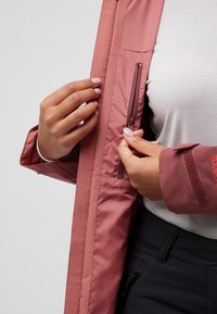 Giacca impermeabile rosa con tasca interna con zip, polsini regolabili con velcro e texture liscia e leggera. Camicia grigia sotto.