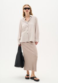 Light beige linen button-up shirt with long sleeves, paired with a ribbed, light pink midi skirt. Model holds a black leather tote bag. Black sandals.
