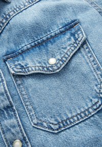Tissu en denim bleu clair présentant une poche poitrine avant avec un rabat courbé et un bouton-pression blanc. Couture visible le long des bords.