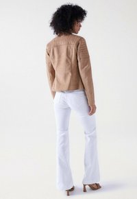 Veste de motard en suede beige avec des détails matelassés sur les épaules, assortie à un jean évasé blanc et des sandales à talons beiges. Vue de derrière.