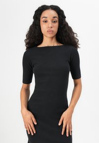 Black ribbed dress with short sleeves, square neckline, fitted silhouette, and smooth texture. Model stands with hands on hips against a light background.
