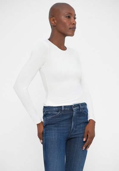 Donna con testa rasata che indossa una camicia bianca attillata a maniche lunghe e jeans blu, in piedi con le mani vicino alle tasche su uno sfondo semplice.